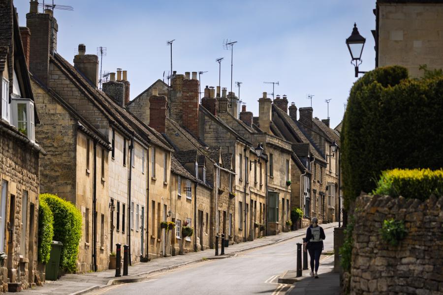 Cotswold Gardens B&B - Wonderful Winchcombe A narrow, gently curving street lined with closely packed stone houses featuring sloped roofs, chimney stacks, and small windows. The buildings vary in warm neutral tones like beige, cream, and light brown, with some greenery climbing walls and hedges along the edges. A single pedestrian walks along the pavement on the right side of the road. A low stone wall and neatly trimmed bush appear in the foreground, while traditional street lamps rise above the homes. The sky overhead is pale and clear, casting soft daylight across the quiet scene in Winchcombe.