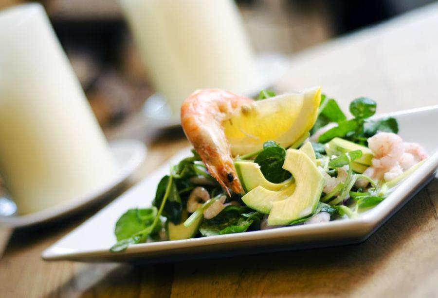 Cotswold Gardens B&B - Wonderful Winchcombe White rectangular plate holding a composed salad with sliced avocado, leafy greens, and pieces of seafood, accompanied by a lemon wedge. The dish is placed on a wooden table, with a softly blurred background featuring a white candle and dining setting, creating a warm restaurant atmosphere.