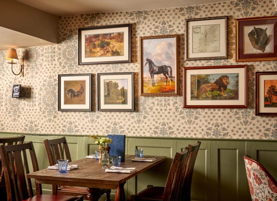 Cotswold Gardens B&B - Wonderful Winchcombe Inside the cosy restaurant at The Lion Inn. A wooden table set for four, surrounded by dark wooden chairs. The wall behind features patterned wallpaper and is decorated with a gallery of framed artwork, many depicting horses and animals in various styles and sizes. A small vase of yellow flowers sits on the table alongside menus, plates, and cutlery. Warm, ambient lighting creates an inviting, traditional atmosphere.